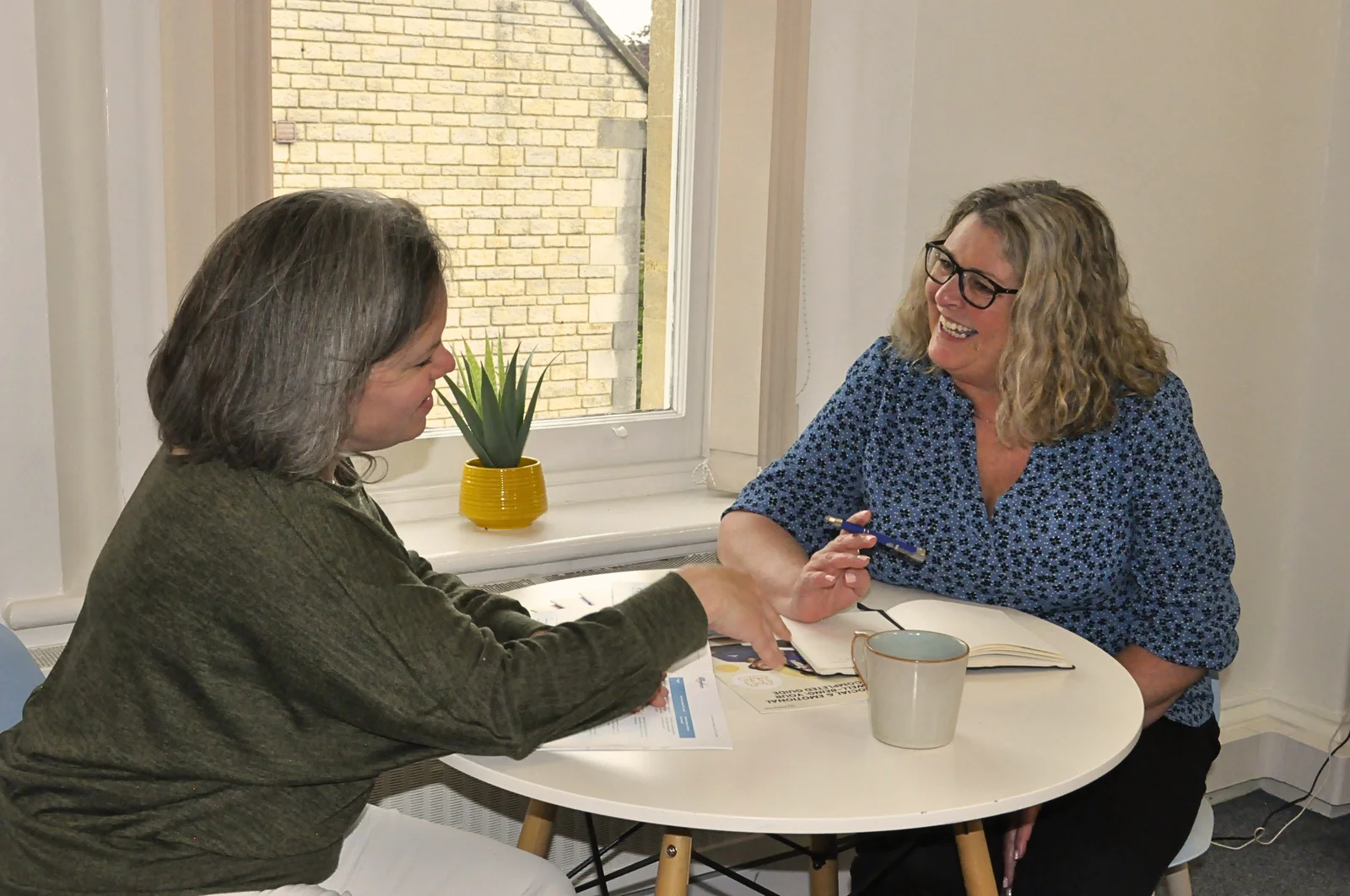 Two women talking together at a table with Programme materials and an open notebook