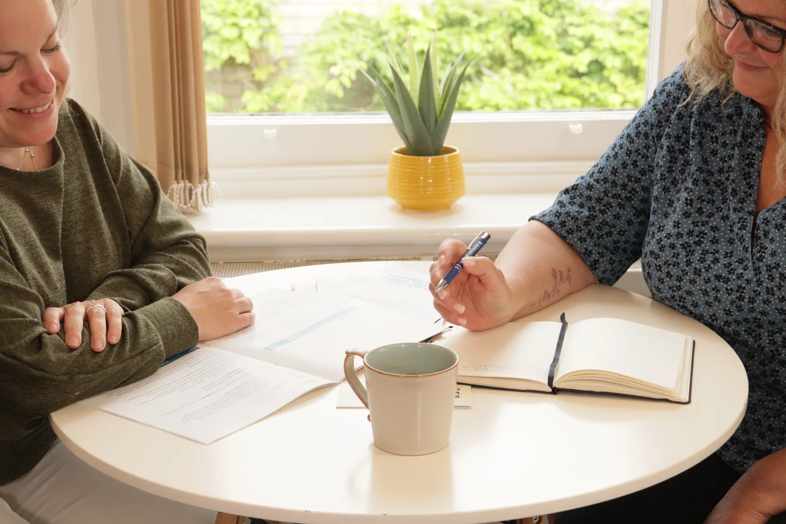 Two colleagues in conversation at a round table by a window, one writing in a notebook beside a mug of tea