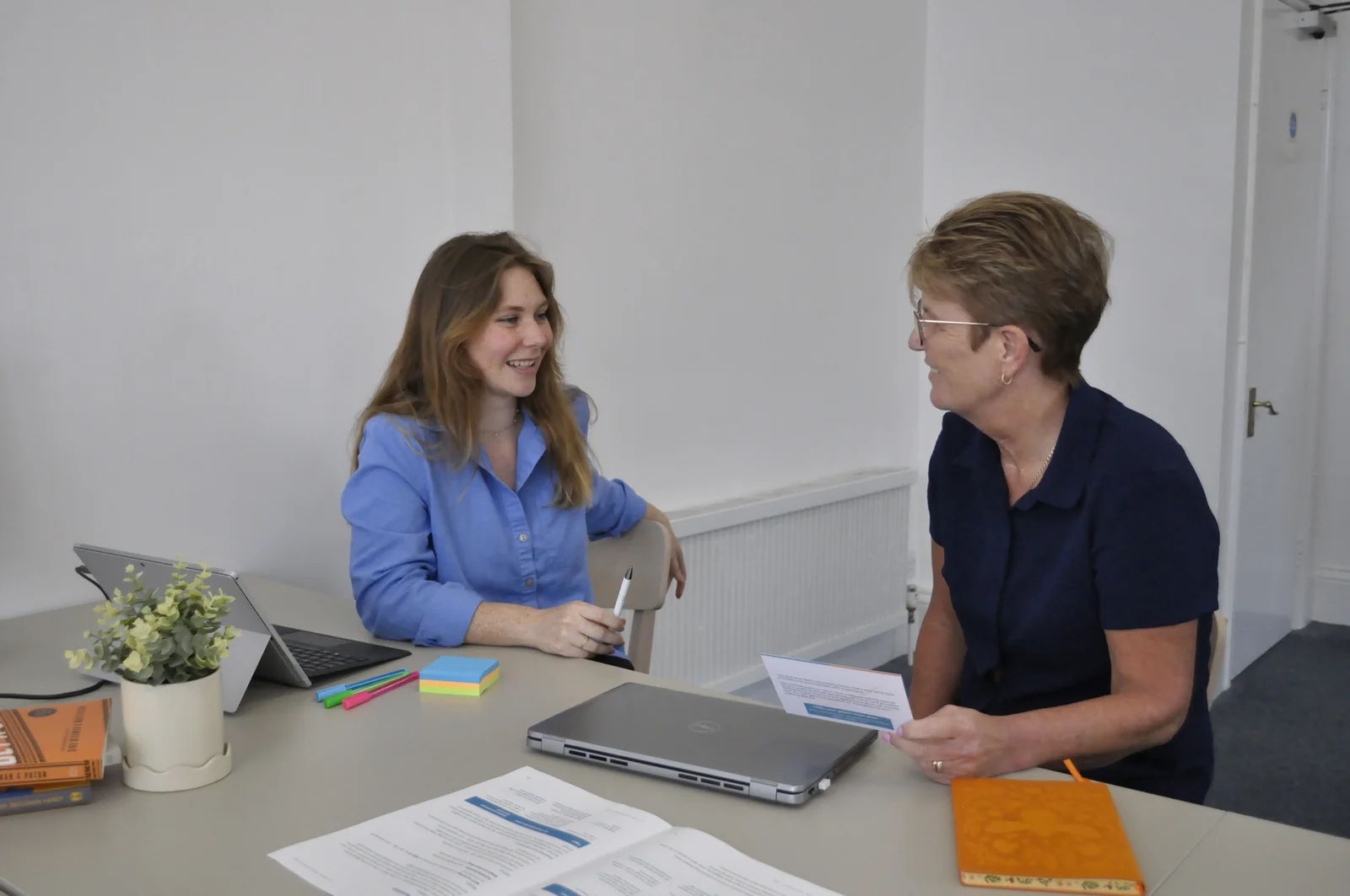 Two women discussing printed Programme materials at a desk with laptops and notebooks