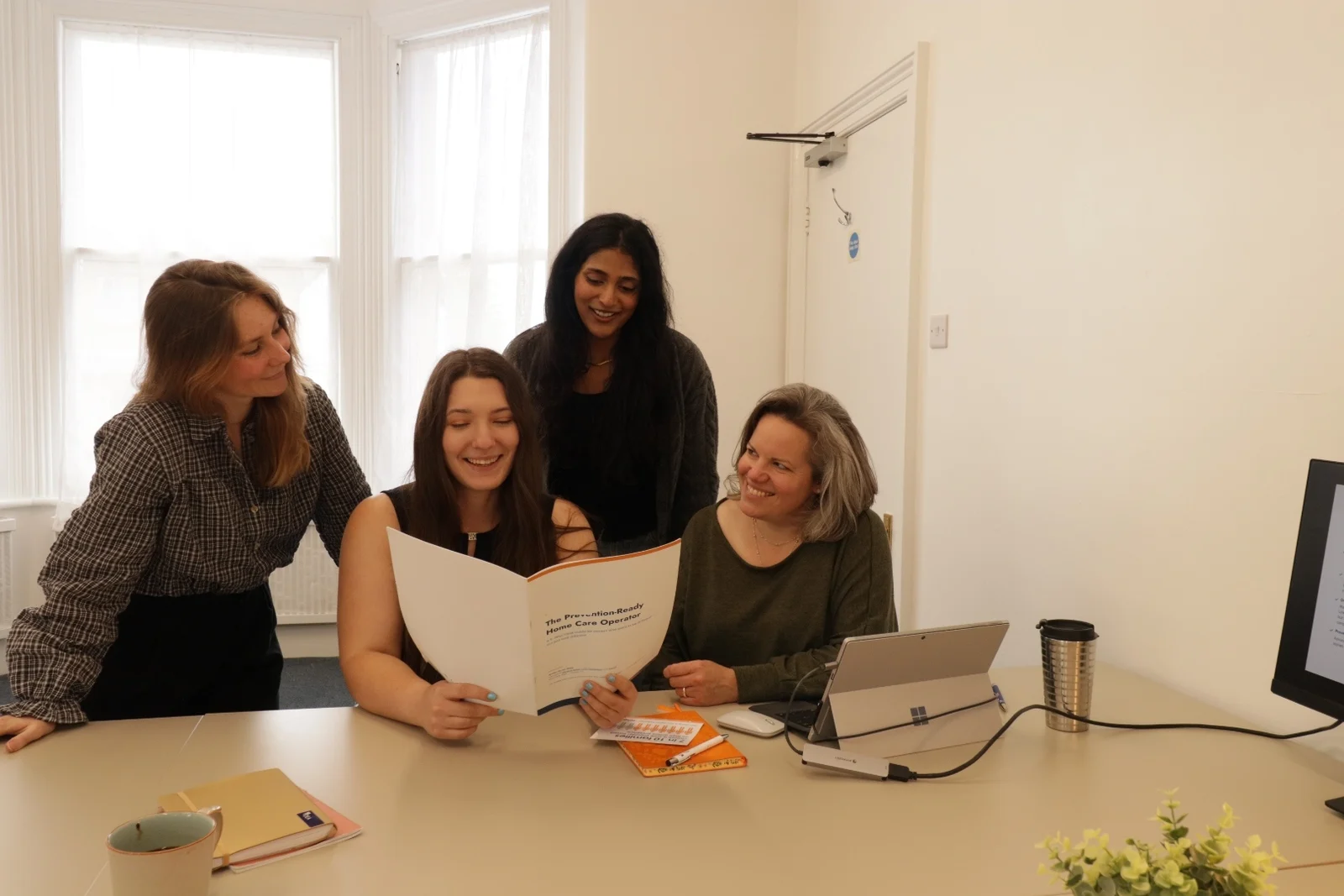 The Five Pillars team standing and sitting around a desk in their Bath office, talking through printed notes and a laptop